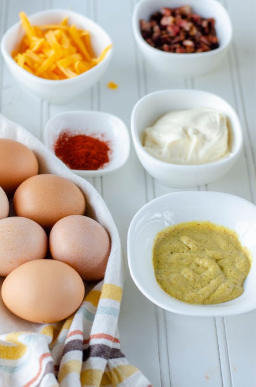 ingredients including hard boiled eggs, mustard, and paprika in white bowls on a white wooden counter