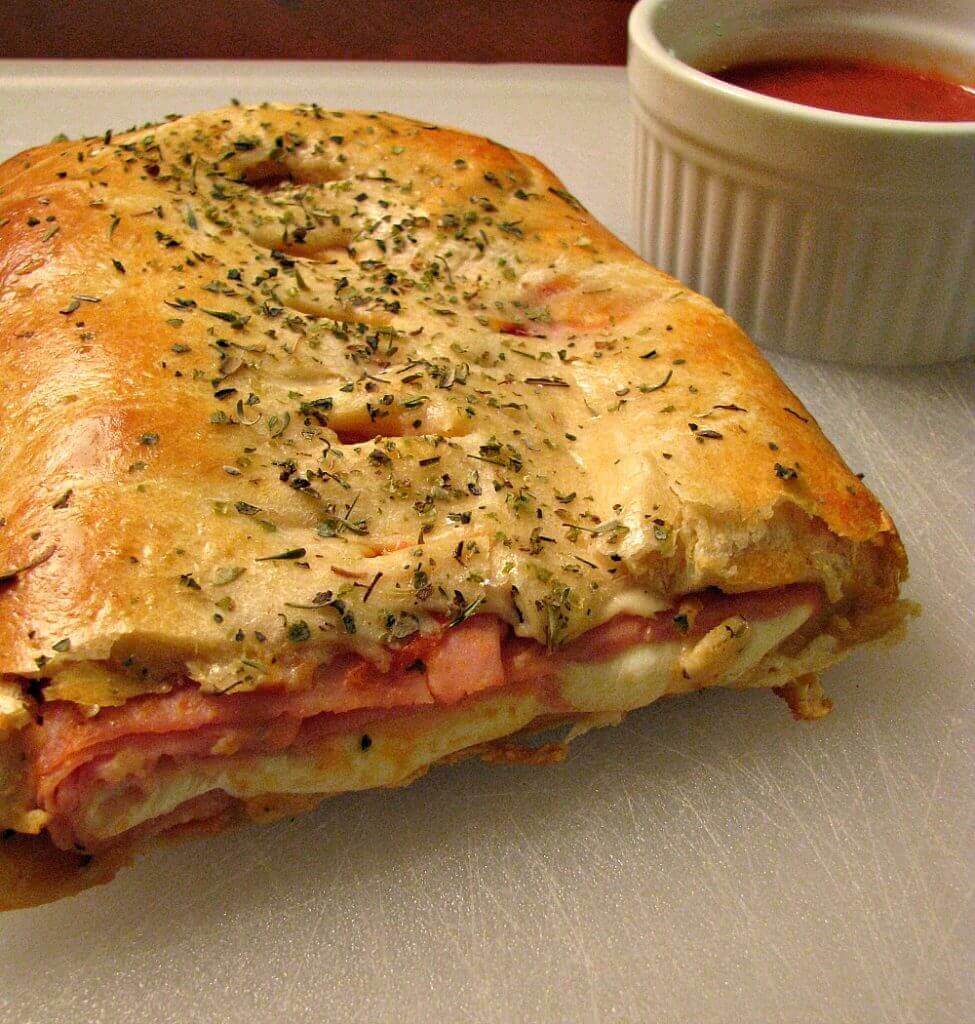 photo of Ham and Cheese Stromboli made with crescent roll dough on a white cutting board with a bowl of marinara sauce. 