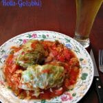 Photo of a plate of two stuffed cabbage rolls with sauce on a flower trimmed white plate next to a glass of beer sitting on a wood table