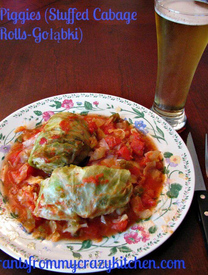 Photo of a plate of two stuffed cabbage rolls with sauce on a flower trimmed white plate next to a glass of beer sitting on a wood table