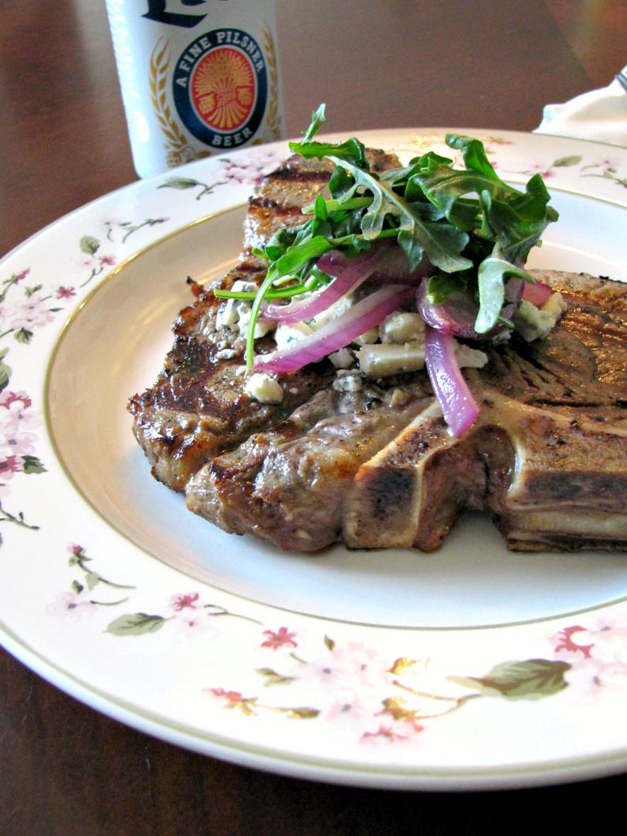 photo of a plate with beer marinated grilled porterhouse steak topped with arugula and blue cheese next to a can of beer.