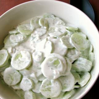 Photo of Creamy Cucumber Salad in a large white bowl on a wood table next to a black serving spoon