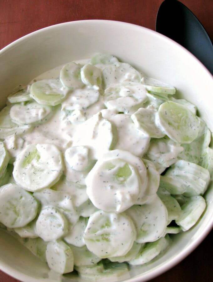Photo of Creamy Cucumber Salad in a large white bowl on a wood table next to a black serving spoon