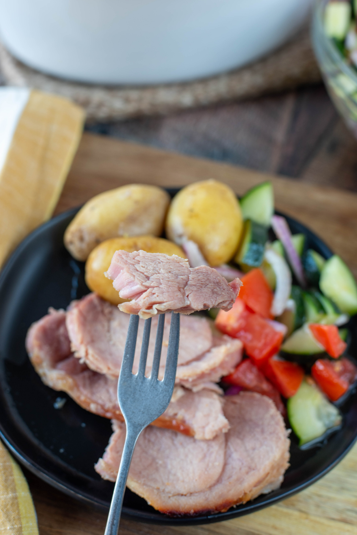 a bite size piece of ham on a fork held above a plate of ham potatoes and salad