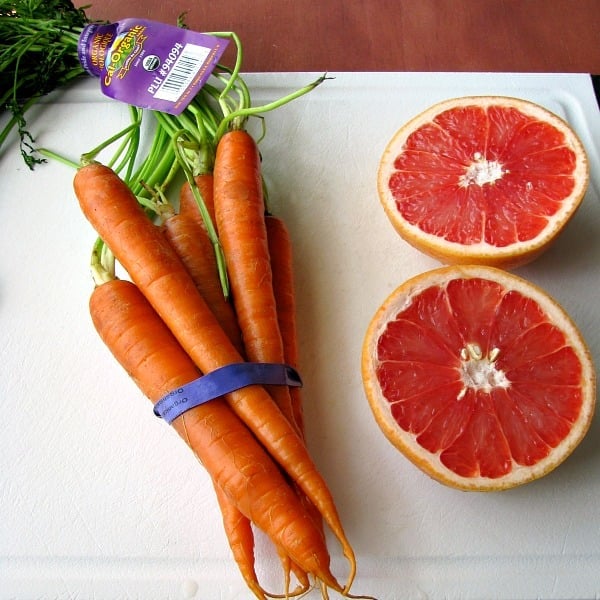 Photo of Cal-Organic Carrots wrapped together with a blue rubber band next to cut open red grapefruit on a white cutting board 