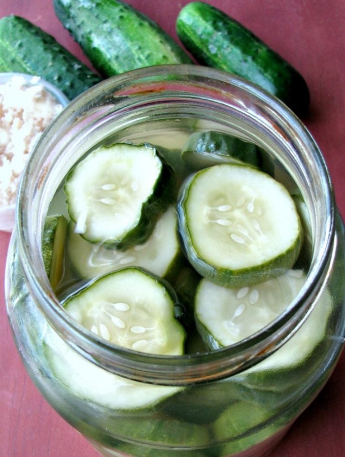 Photo of Sweet Horseradish Pickles in a glass jar on a wood table next to pickling cucumbers and a small bowl of chopped horseradish
