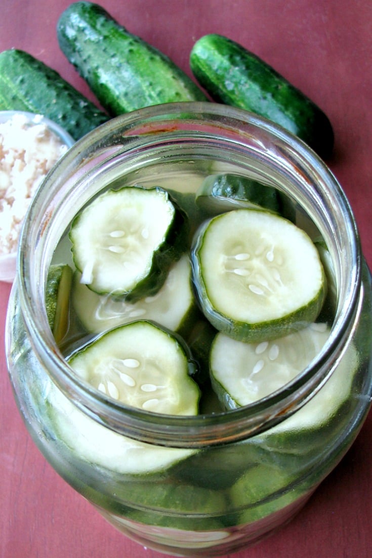 Photo of Sweet Horseradish Pickles in a glass jar on a wood table next to pickling cucumbers and a small bowl of chopped horseradish