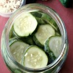 Photo of a glass jar filled with Sweet Horseradish Pickles on a wood table next to a small bowl of chopped horseradish and pickling cucmbers