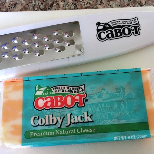 Photo of a package of Cabot Colby Jack cheese on a white cutting board next to a cheese grater with the Cabot logo on it