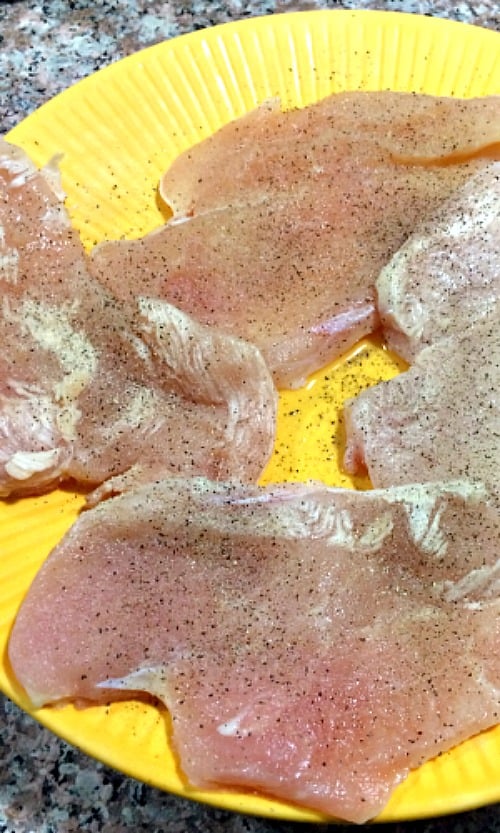 Photo of raw chicken breasts seasoned with salt, pepper, and garlic powder on a yellow plate 