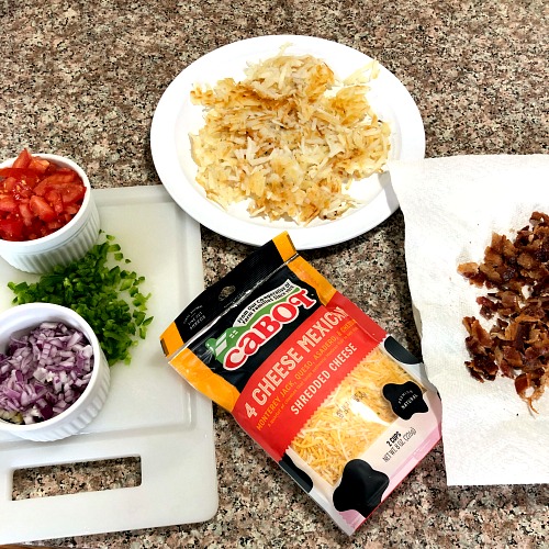 Photo of ingredients for Cheesy Baked Breakfast Tacos including fried hash browns on a white plate crumbled bacon on a white paper plate a package of Cabot's shredded Mexican cheese blend and diced tomatoes onions and jalapenos on a white cutting board 