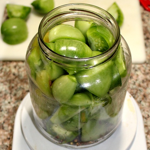 photo of sliced green tomatoes packed into a jar in front of a cutting board with more green tomatoes