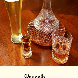 photo of a shot glass of Krupnik (honey spiced liqueur) in a shot glass next to a glass of beer a bottle and a glass on a wood table