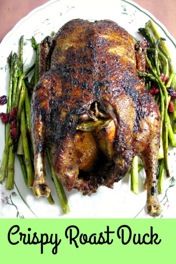 Photo of a whole Crispy Roast Duck on a white flower trimmed platter with roasted asparagus and cranberries on the sides
